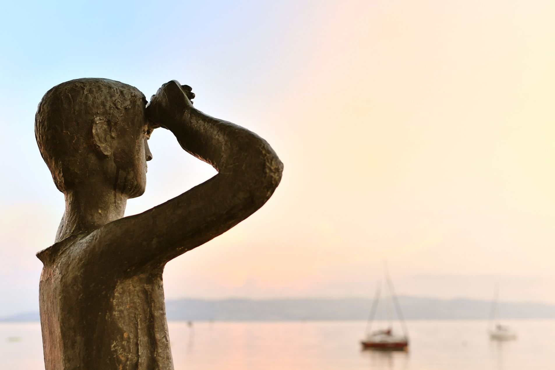 Das Denkmal des lieben Augustin in Wasserburg. Im Hintergrund ist verschwommen der Bodensee mit Segelboten zu sehen.