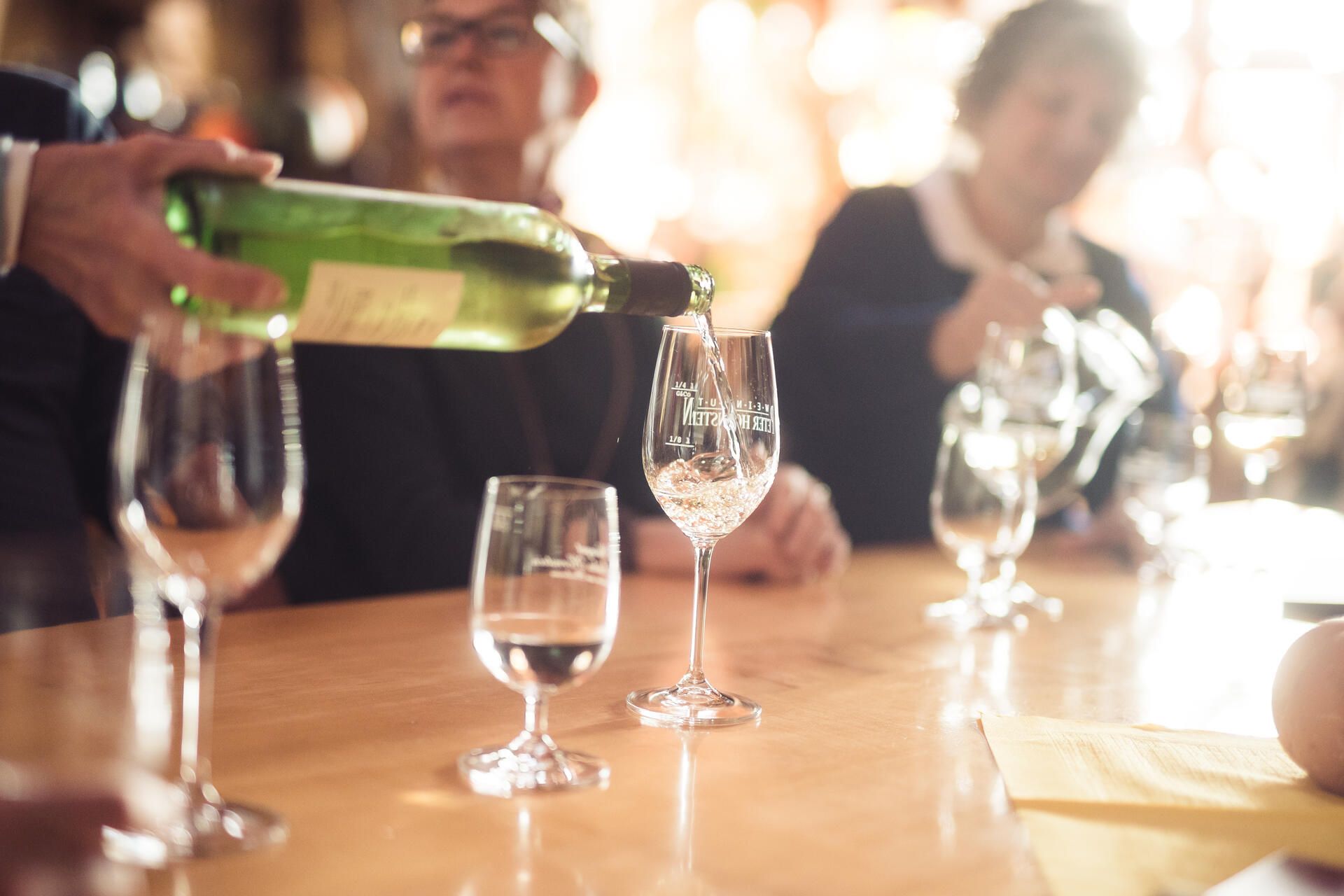 Wein wird in ein Weinglas eingefüllt. Im Hintergrund sieht man verschwommen Leute die an einem Tisch sitzen.