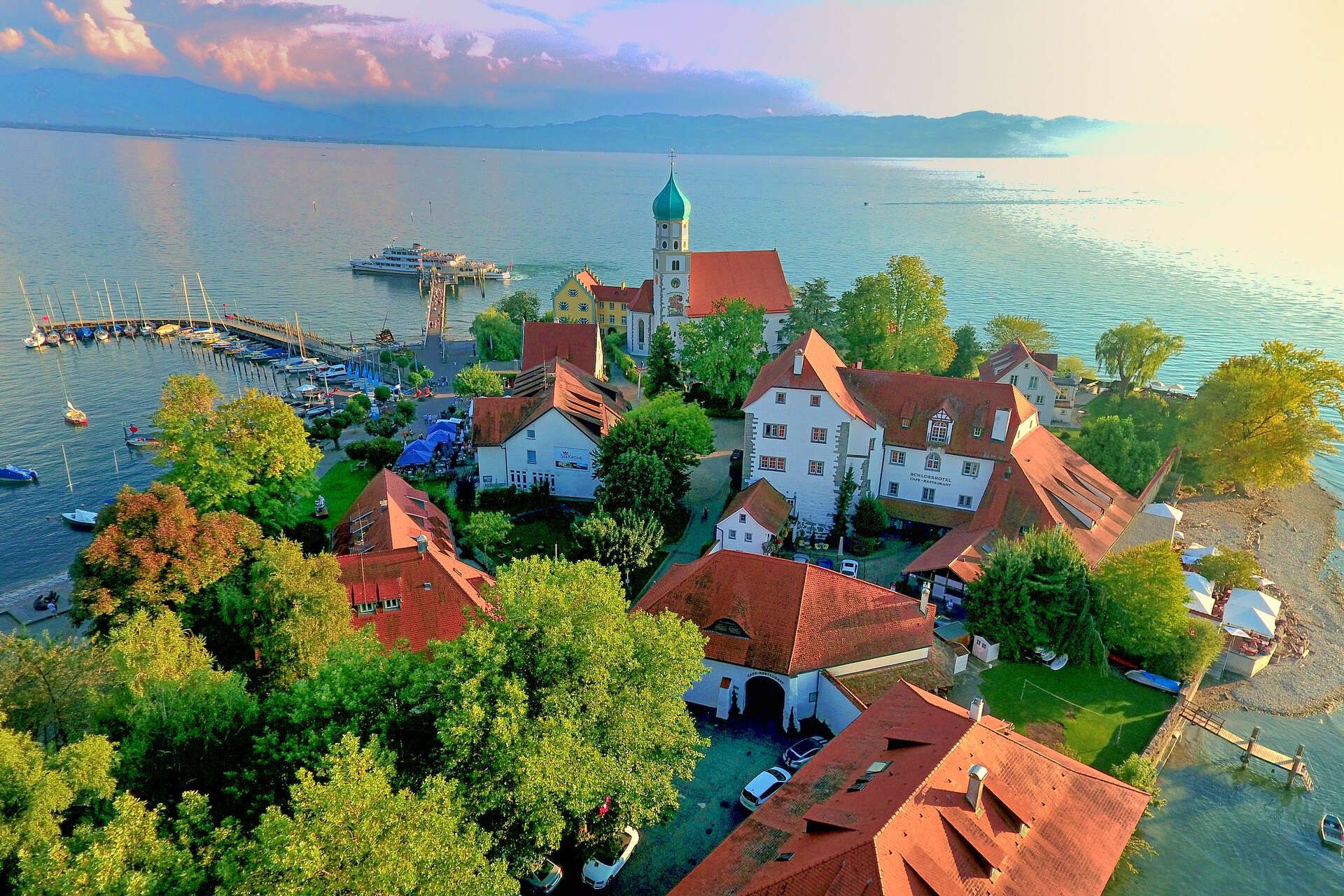 Luftaufnahme der Halbinsel in Wasserburg. Umringt vom Bodensee, im Hintergrund ist die Bergwelt zu sehen.