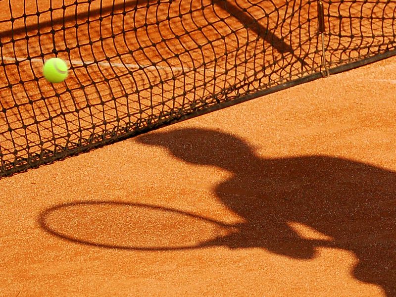Tennisfeld mit Netz ein Tennisball wird ins Netz gespielt. Es ist ein Schatten von einer Person mit einem Tennisschläger zu sehen.