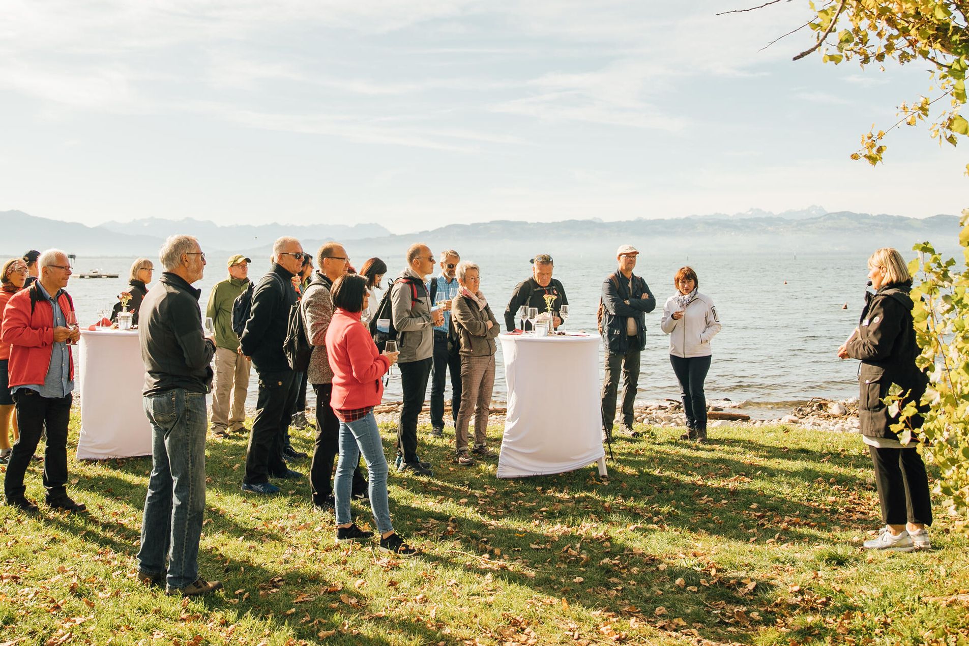 Eine Gästeführerin erklärt einer Gruppe was. Die Gruppe verköstigt Wein. Im Hintergrund ist der Bodensee und die Bergwelt zu sehen.