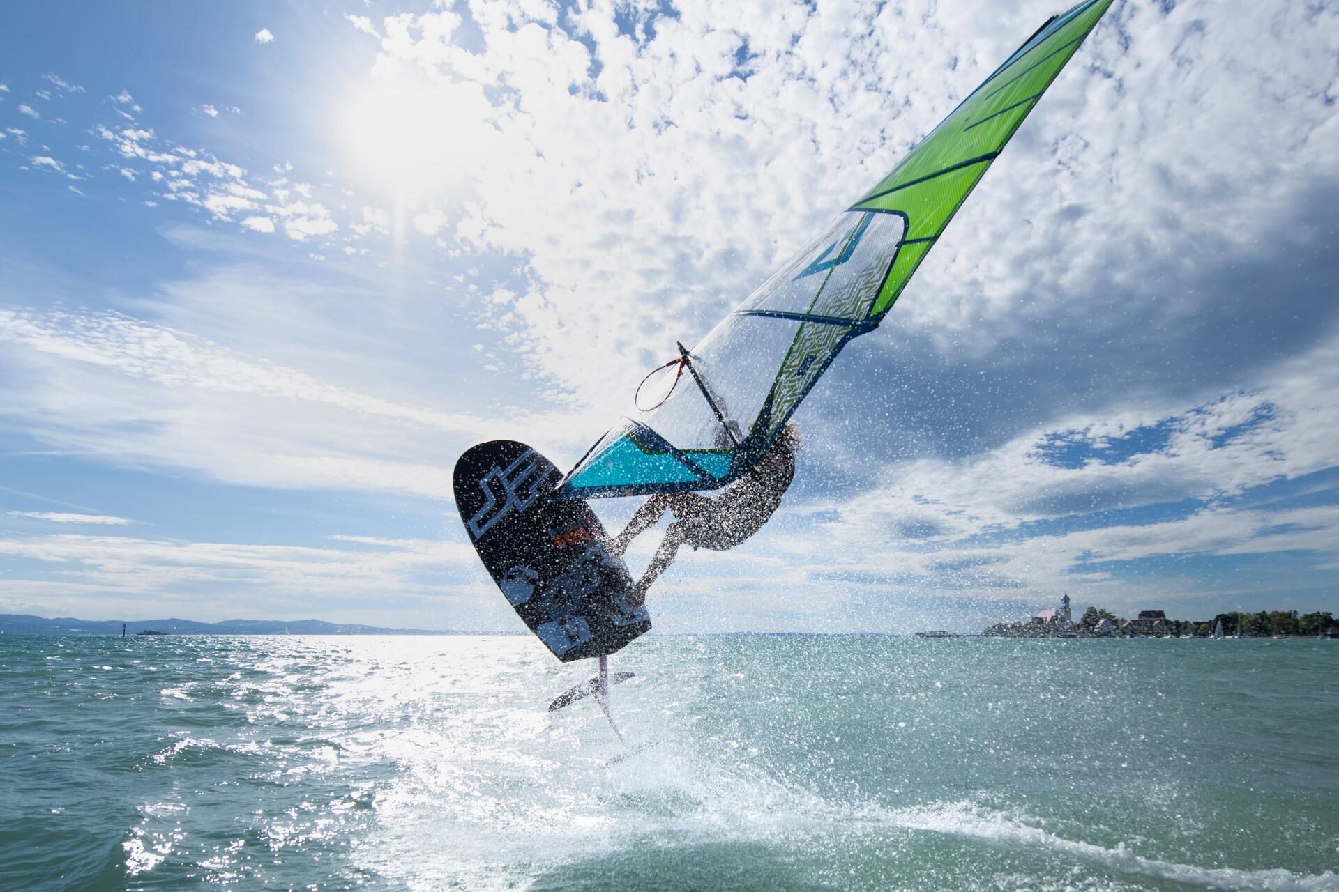 Ein Windsurfer auf dem Bodensee jumpt in höhe. Im Hintergrund ist die Halbinsel in Wasserburg zu sehen.