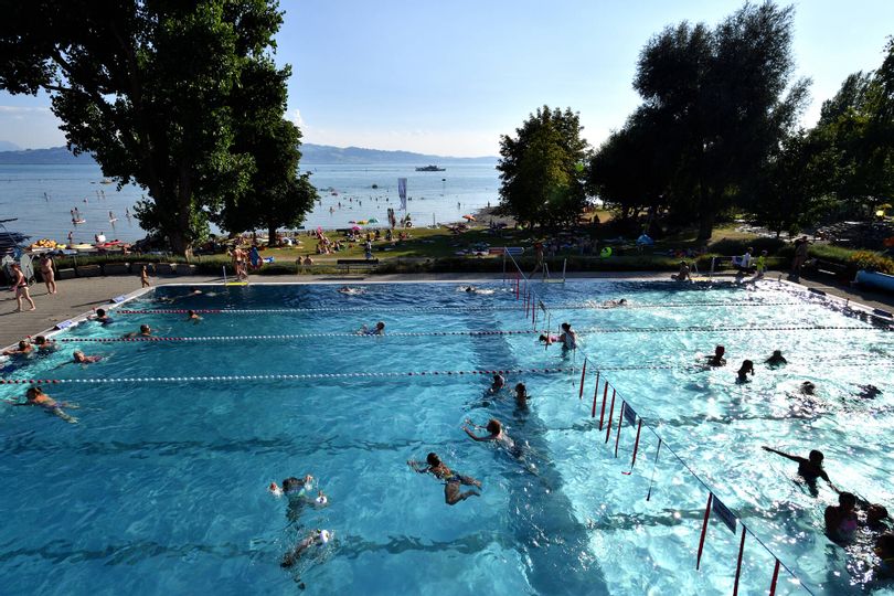 Liegewiese, Schwimmbecken und Seezugang im Freibad Aquamarin in Wasserburg. Im Hintergrund ist der See und die Bergwelt zu sehen.