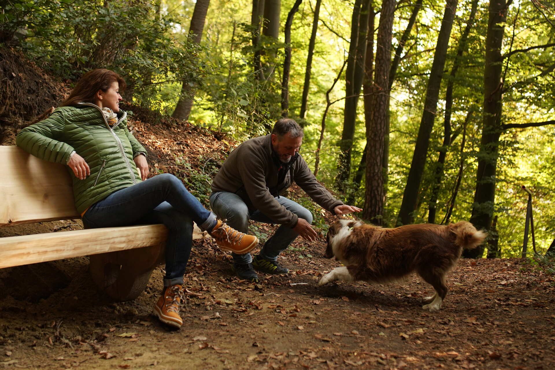 Eine Frau und ein Mann sitzen auf einer Bank im Wald und spielen mit einem Hund.