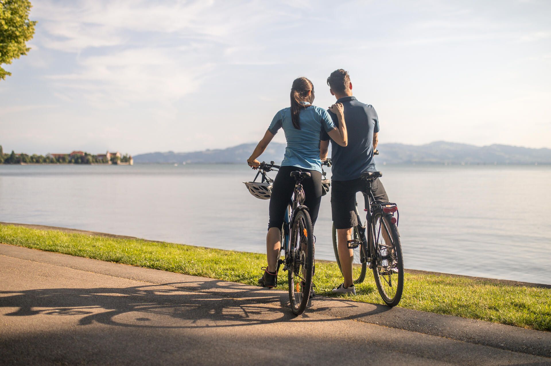 Ein Pärchen fährt auf Panorama Radrunde am Lindauer Bodensee Fahrrad. Der WEg liegt direkt am Bodensee. Im Hintergrund ist die Bergwelt zu sehen.