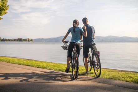 Ein Pärchen fährt auf Panorama Radrunde am Lindauer Bodensee Fahrrad. Der WEg liegt direkt am Bodensee. Im Hintergrund ist die Bergwelt zu sehen.