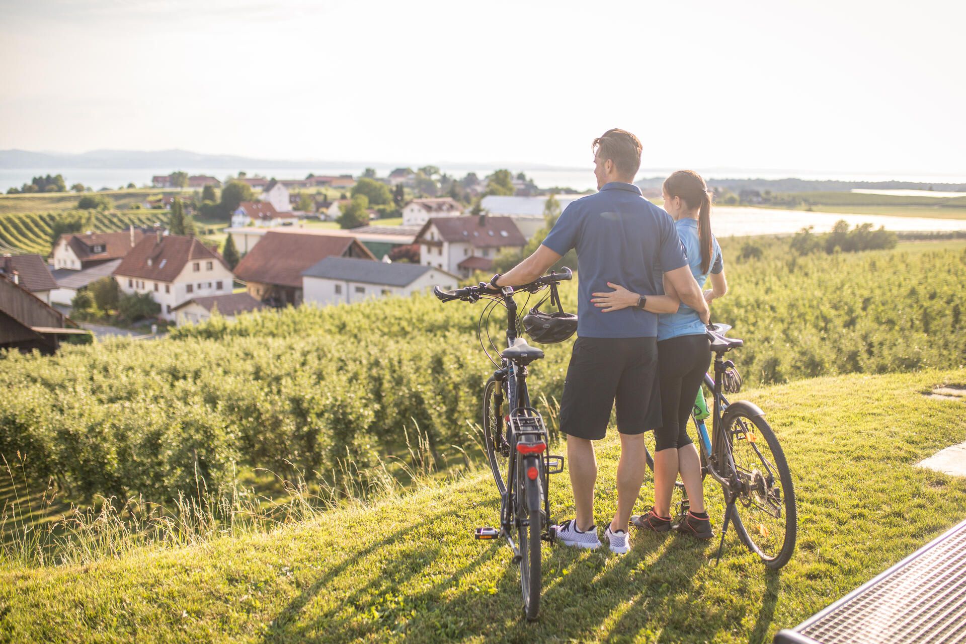 Zwei Radfahrer genießen die Aussicht an der Antoniuskapelle in Selmnau. Im Hintegrund ist der See und die Bergwelt zu sehen.