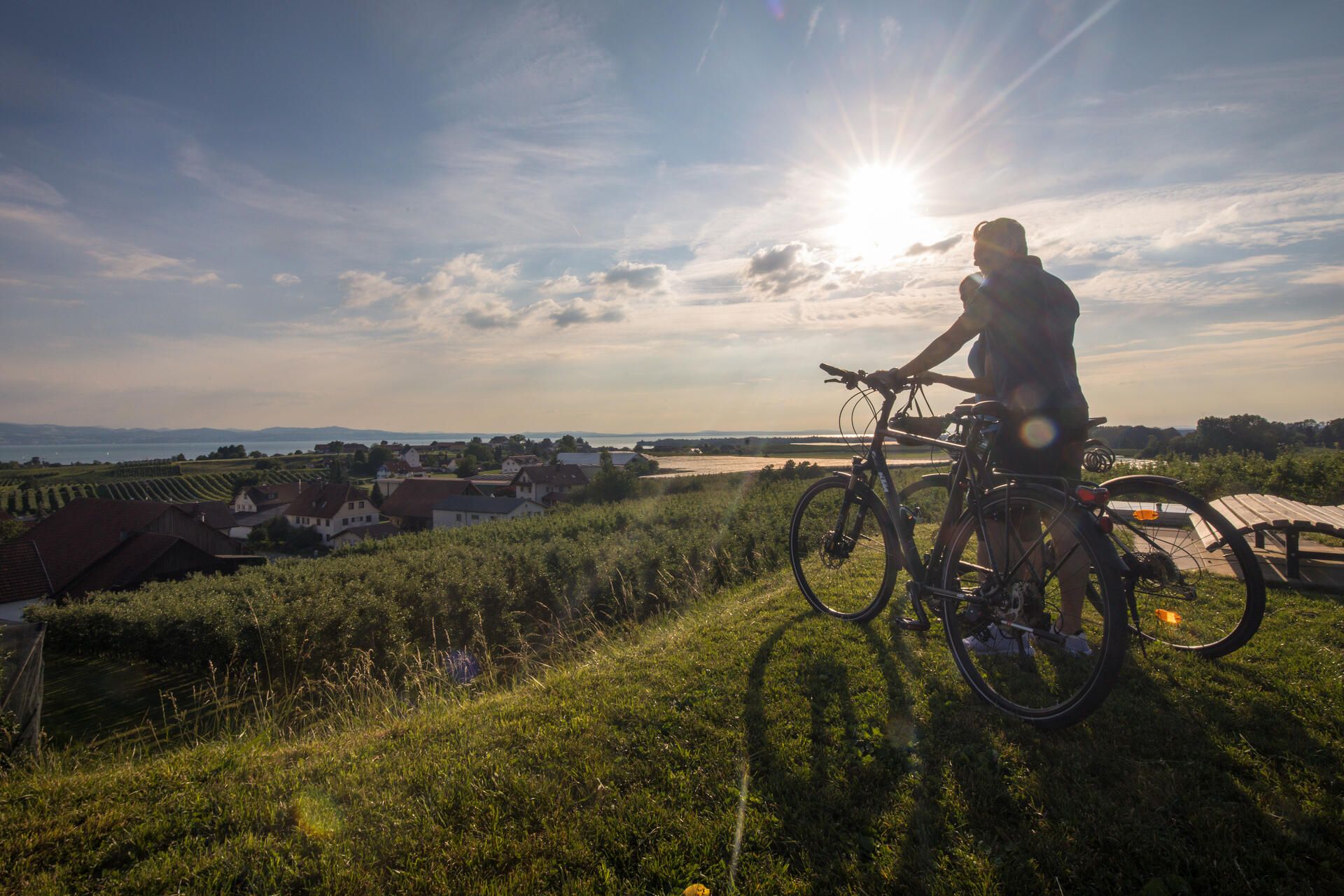 Zwei Radfahrer genießen die Aussicht an der Antoniuskapelle in Selmnau. Im Hintegrund ist der See und die Bergwelt zu sehen.