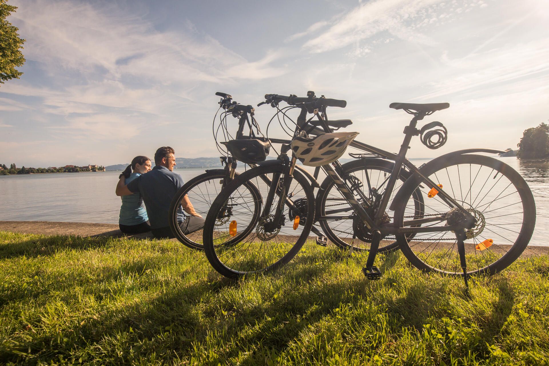 Zwei Fahrräder stehen in der Wiese vor einem Mäuerchen am Bodensee. Im Hintergrund sieht man ein Pärchen auf dem Mäuerchen sitzen sowie den See und die Bergwelt.