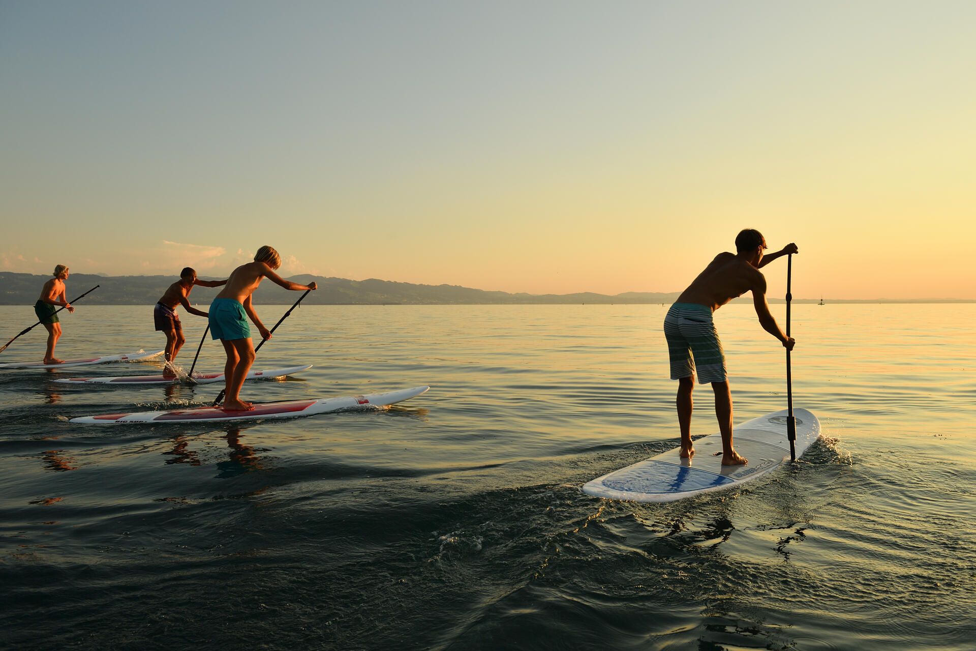 Vier Jugendliche auf ihren SUPs auf dem Bodensee. Im Hintergrund die Bergwelt und ein leichter Sonnenuntergang.
