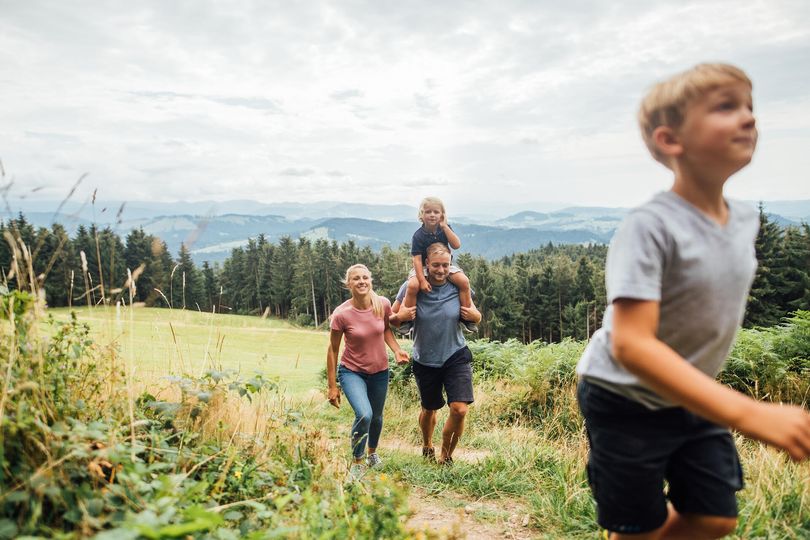 Eine Familie die auf einem Wanderweg inmitten von Grün läuft. Im Hintergrund ist die Bergwelt zu sehen.