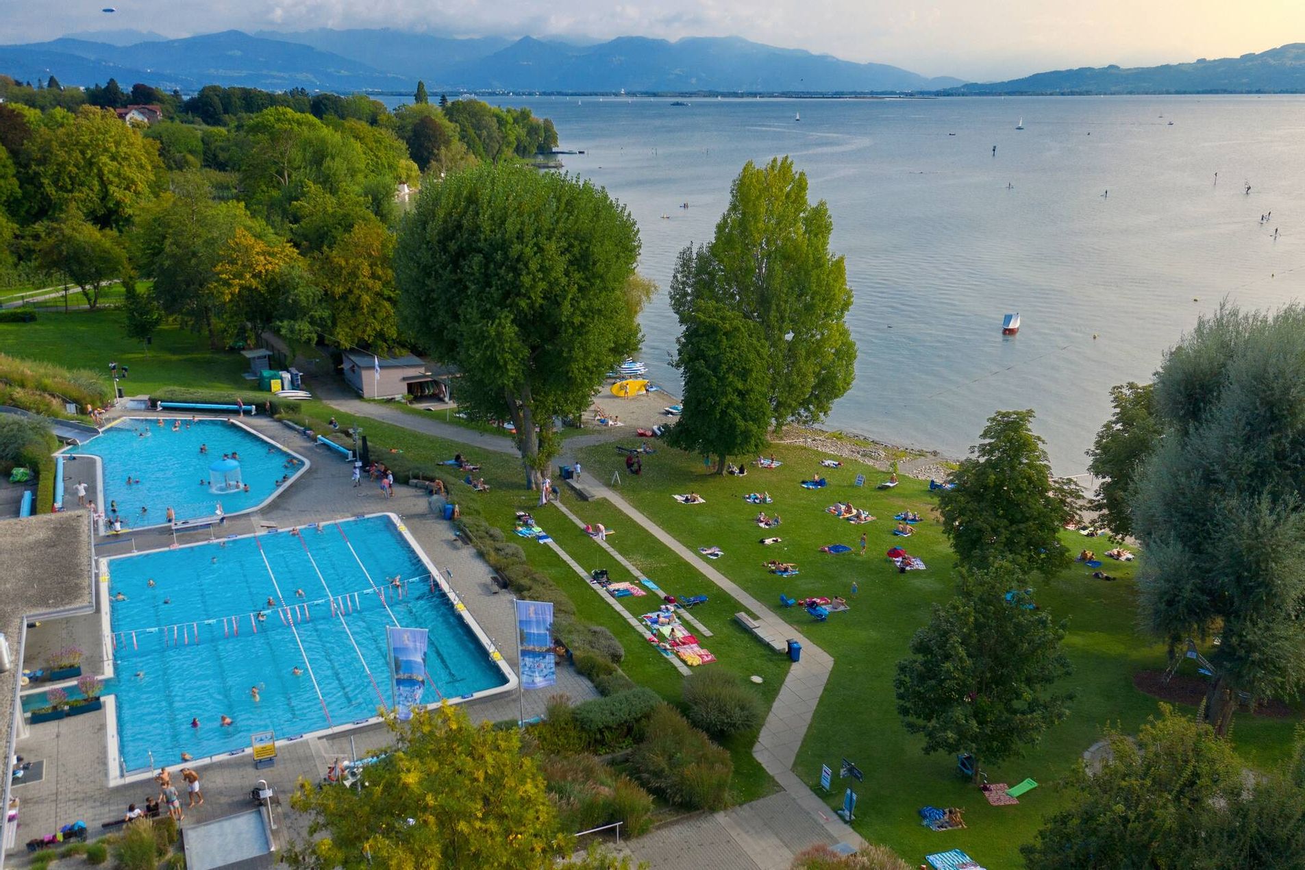 Luftaufnahme Freibad Aquamarin mit Beckenbereich, Liegewiese und dem Seezugang. Im Hintergrund ist die Bergwelt zu sehen.