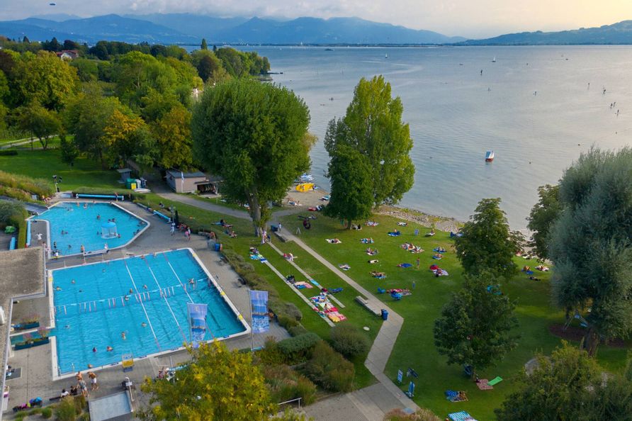 Luftaufnahme Freibad Aquamarin mit Beckenbereich, Liegewiese und dem Seezugang. Im Hintergrund ist die Bergwelt zu sehen.