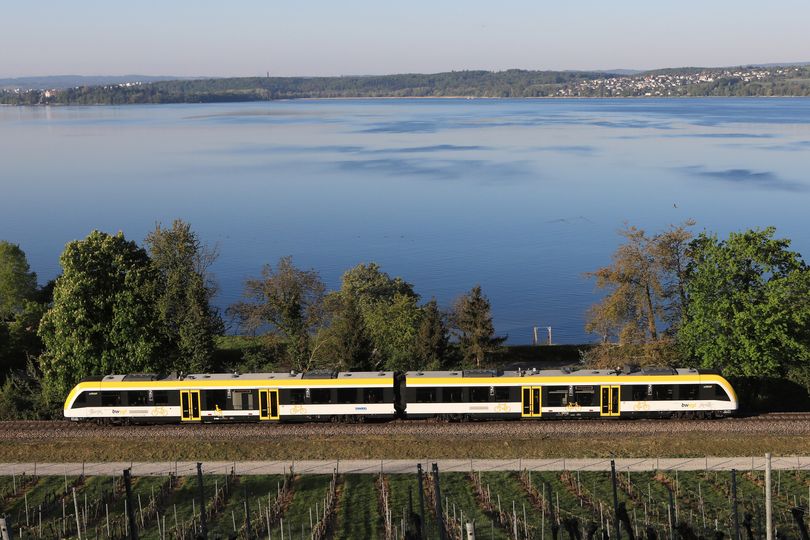 Luftaufnahme Zug der Bodenseegürtelbahn, im Hintergrund ist der Bodensee und Bergwelt zu sehen.