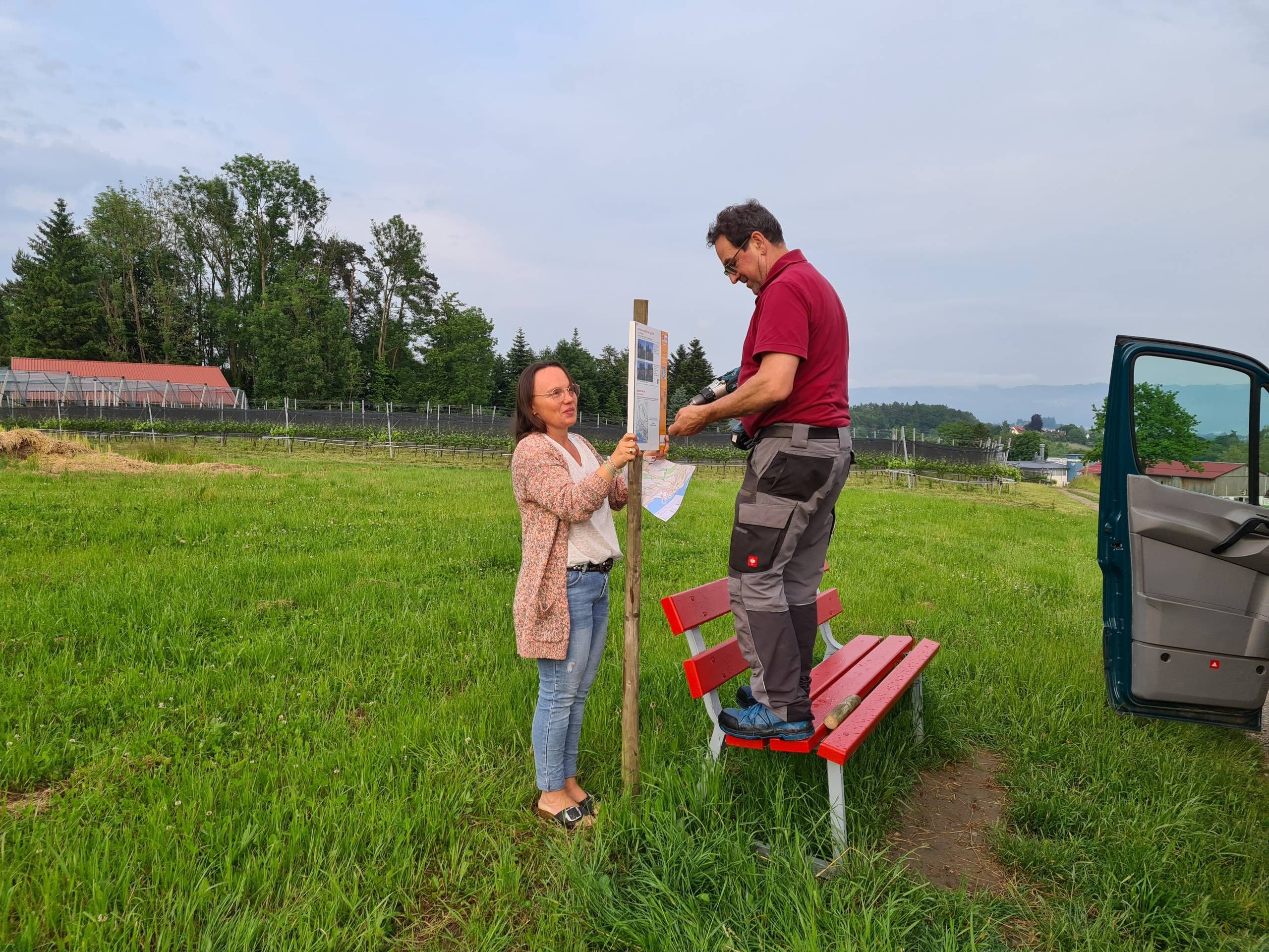 Anbringung der Tafel Trimm.dich.Pfad Wasserburg am Weg des Pfades inmitten von Grün. Im Hintergrund sieht man ein Wald.