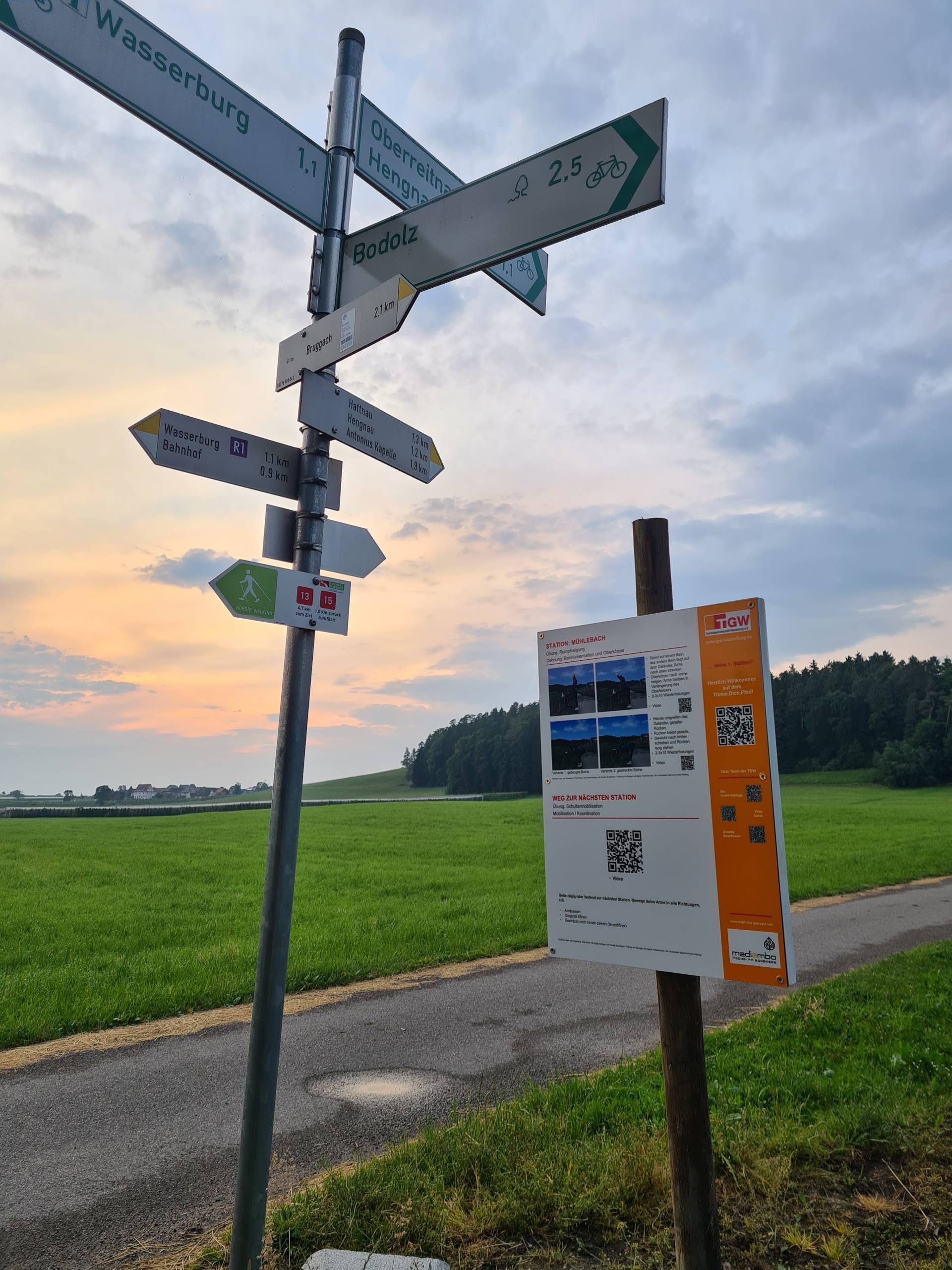 Tafel und Wanderwegbeschreibung des Trimm.dich.Pfad Wasserburg. Im Hintergrund ist grüne Wiese und ein Wald zu sehen.