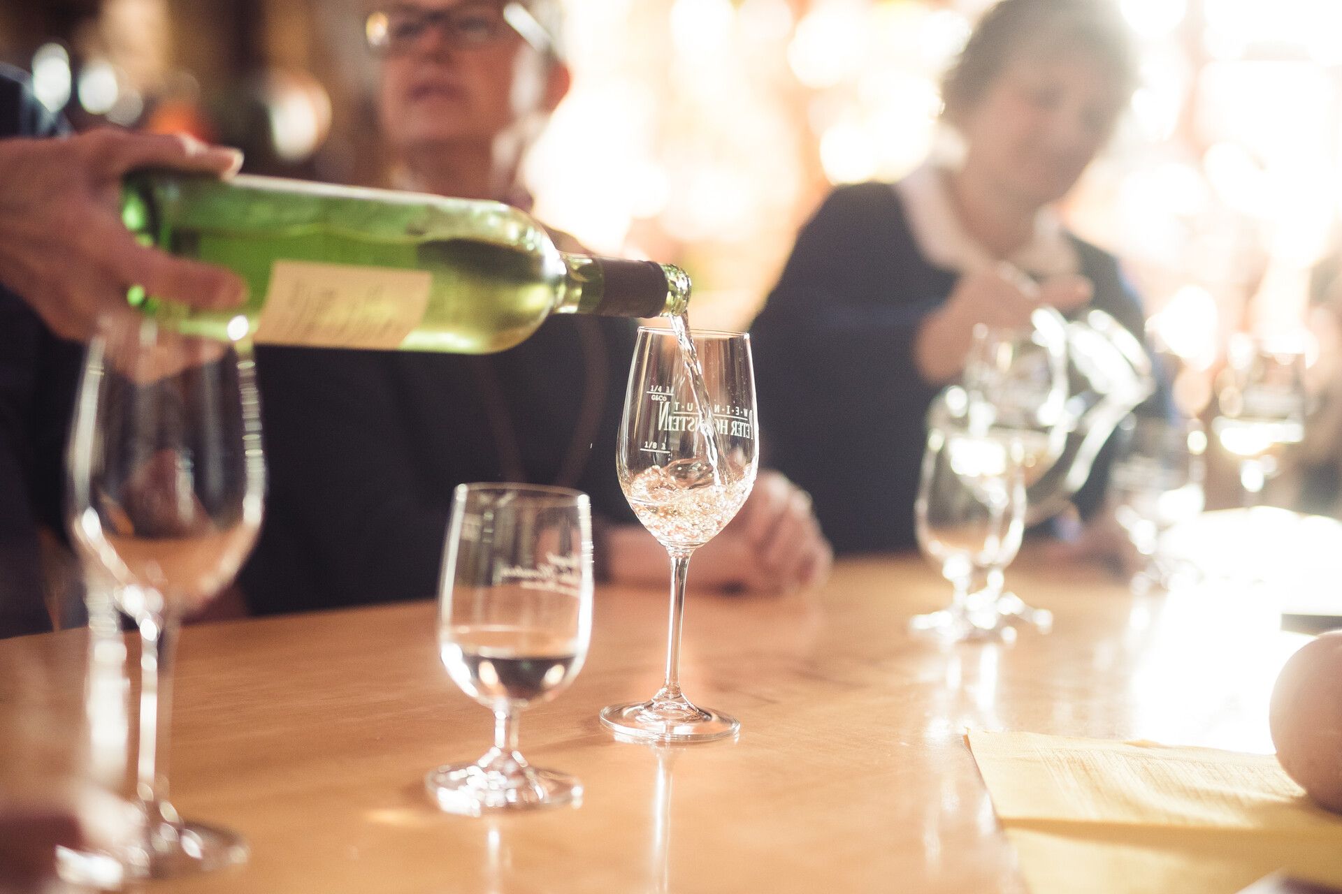 Wein wird in ein Weinglas eingefüllt. Im Hintergrund sieht man verschwommen Leute die an einem Tisch sitzen.