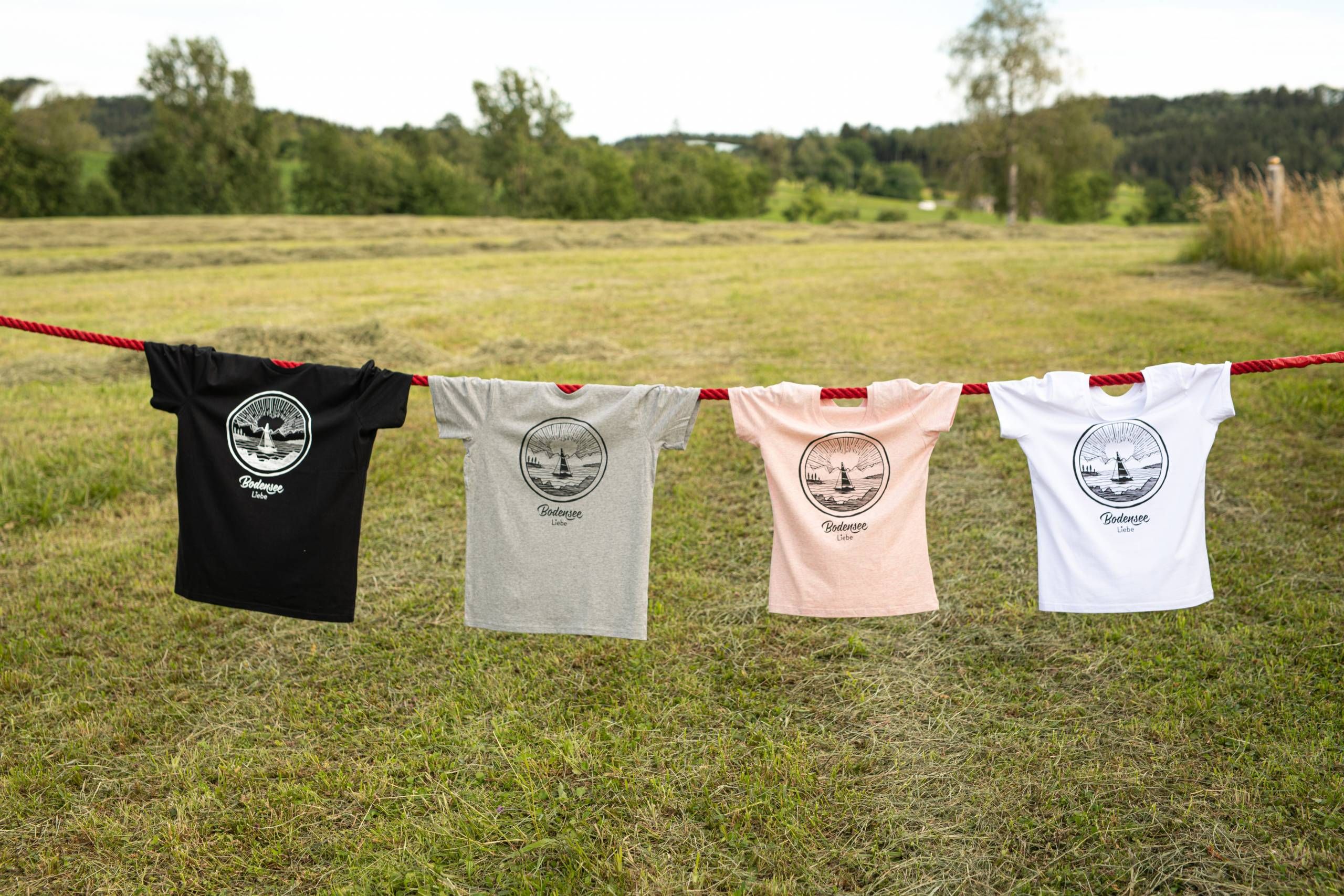 An einer Leine auf einem Feld im Grünen sind mehre T-Shirts vom Bodenseeliebe aufgehängt.