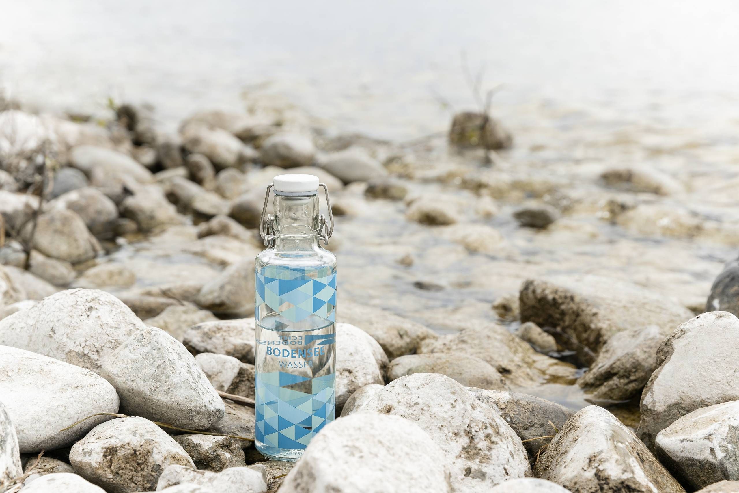 Glasflasche ECHT BODENSEE Trinkflasche steht in mitten von Steinen am Wasser vom Bodensee