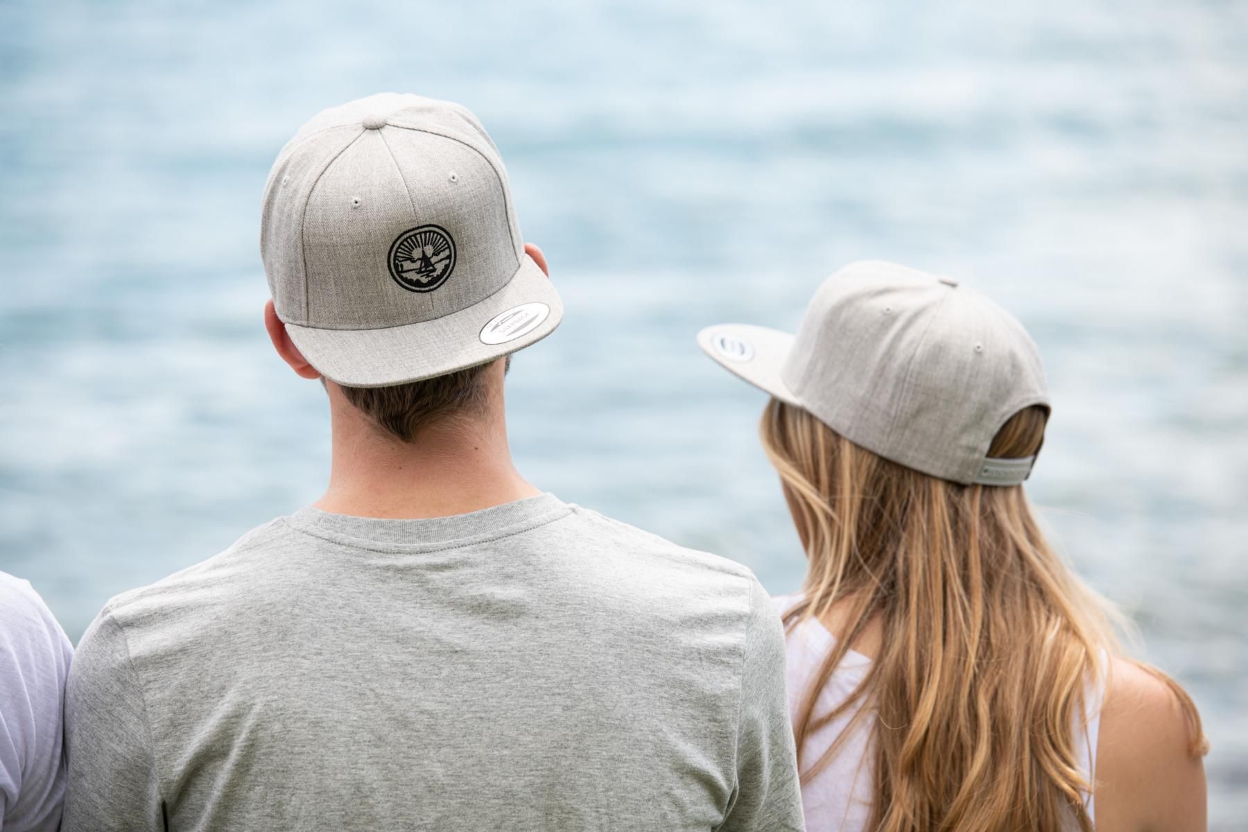 Eine Frau und ein Mann die eine Bodenseeliebe Cap aufhaben. Im Hintergrund ist verschwommen der See zu sehen.