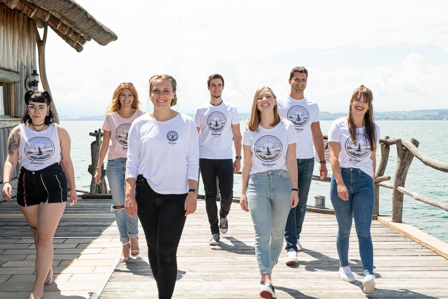Eine Gruppe von Männern und Frauen die ein Oberteil von Bodenseeliebe an haben im Hintergrund ist der Bodensee zu sehen.