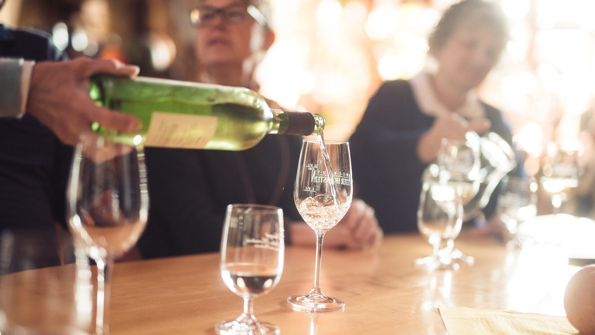 Wein wird in ein Weinglas eingefüllt. Im Hintergrund sieht man verschwommen Leute die an einem Tisch sitzen.