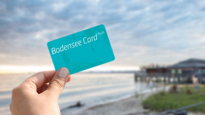 Eine Hand hält die Bodensee Card Plus in Hand im Hintergrund verschwommen der See.