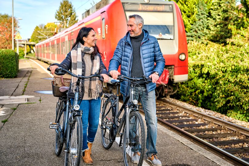 Ankunft eines Zuges am Bahnhof im Vordergrund eine Frau und ein Mann mit ihren Fahrrädern