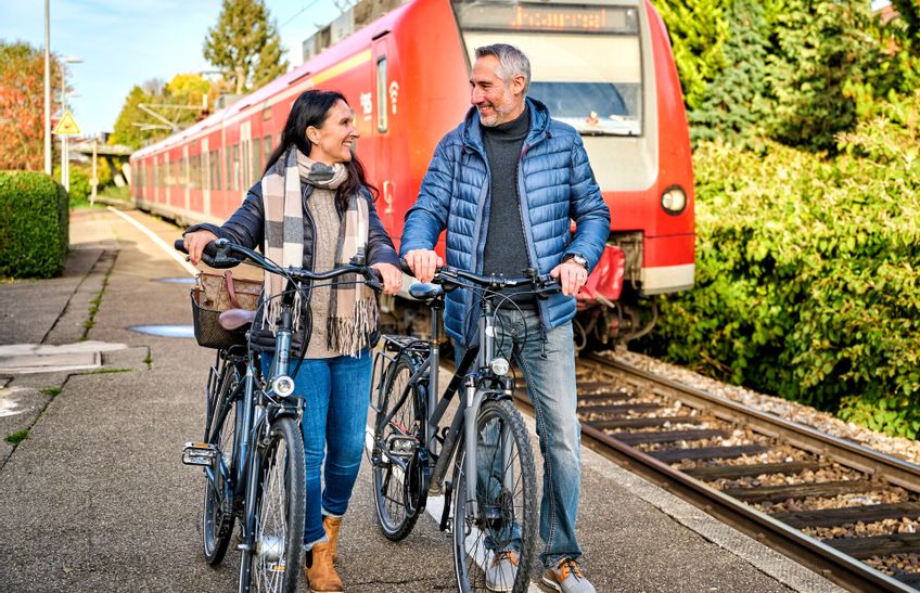 Ankunft eines Zuges am Bahnhof im Vordergrund eine Frau und ein Mann mit ihren Fahrrädern