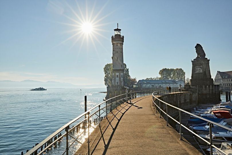 Lindauer Hafen mit Hafeneinfahrt, Leuchtturm, Lindauer Löwe, Löwendenkmal. Im Hintergrund der Bodensee und eine strahlende Sonne.