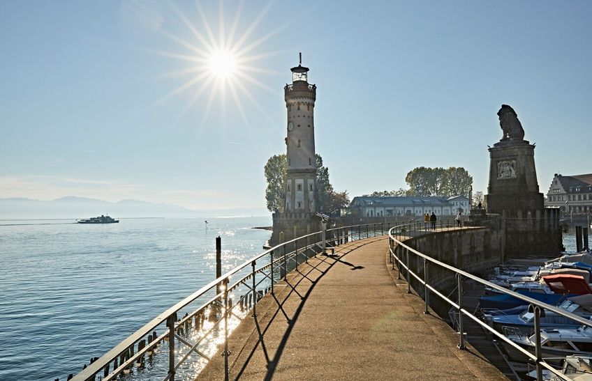 Lindauer Hafen mit Hafeneinfahrt, Leuchtturm, Lindauer Löwe, Löwendenkmal. Im Hintergrund der Bodensee und eine strahlende Sonne.