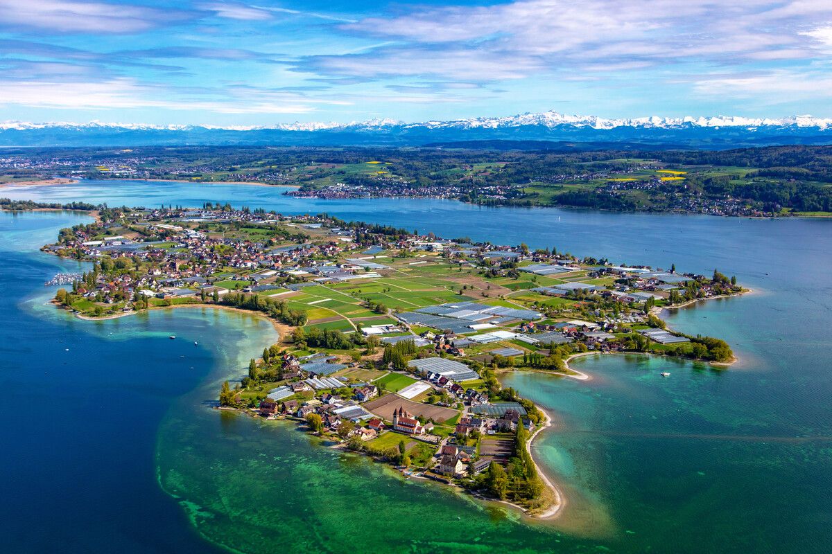 Luftaufnahme der Insel Reichenau drumherum der Bodensee. Im Hintergrund schneebedeckte Berge