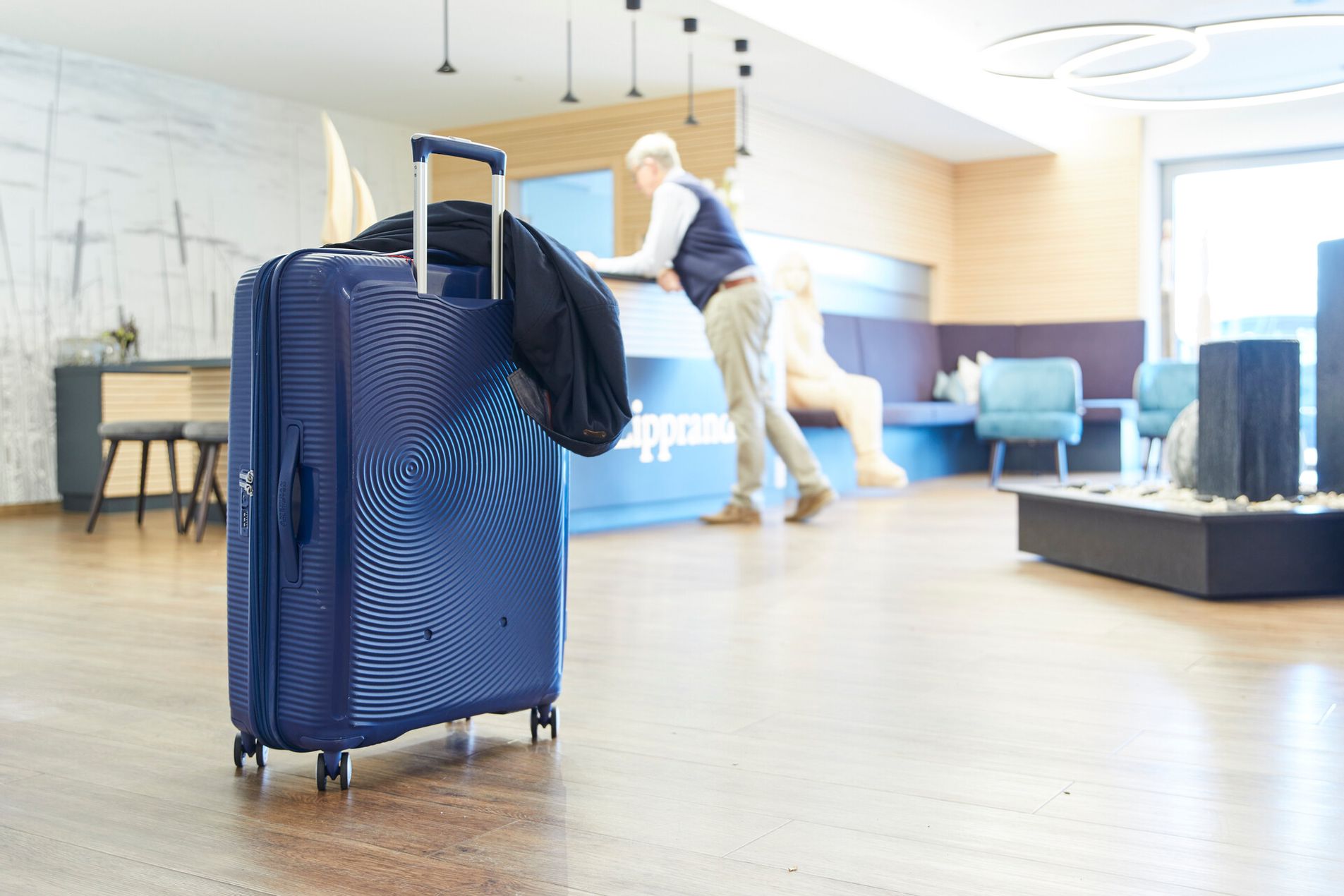 Im Vordergrund steht ein Rollkoffer im Hintergrund ein Mann an der Rezeption