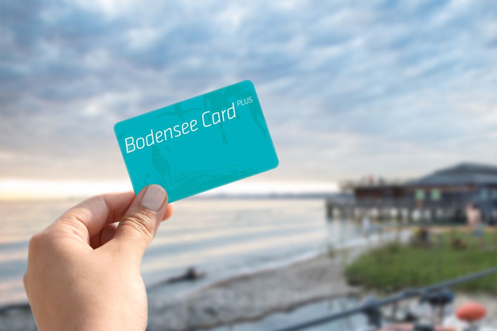 Eine Hand hält die Bodensee Card Plus in Hand im Hintergrund verschwommen der See.