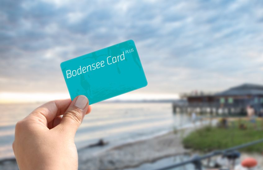 Eine Hand hält die Bodensee Card Plus in Hand im Hintergrund verschwommen der See.