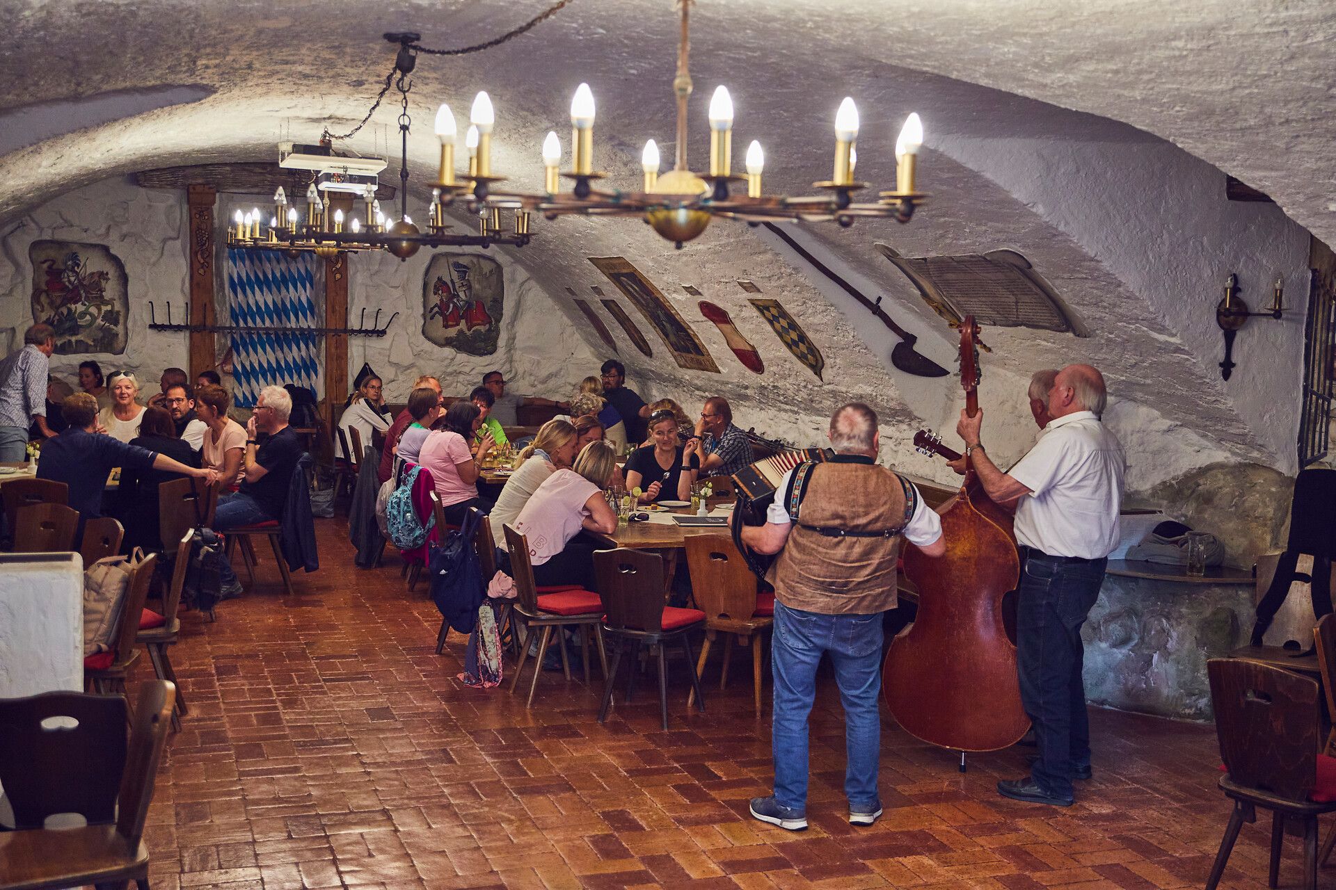 Mehrere Personen sitzen im Schlosskeller vom Schlosshotel Wasserburg an Tischen umrahmt werden sie mit einem Musikalischen Programm des Trio Spontan.