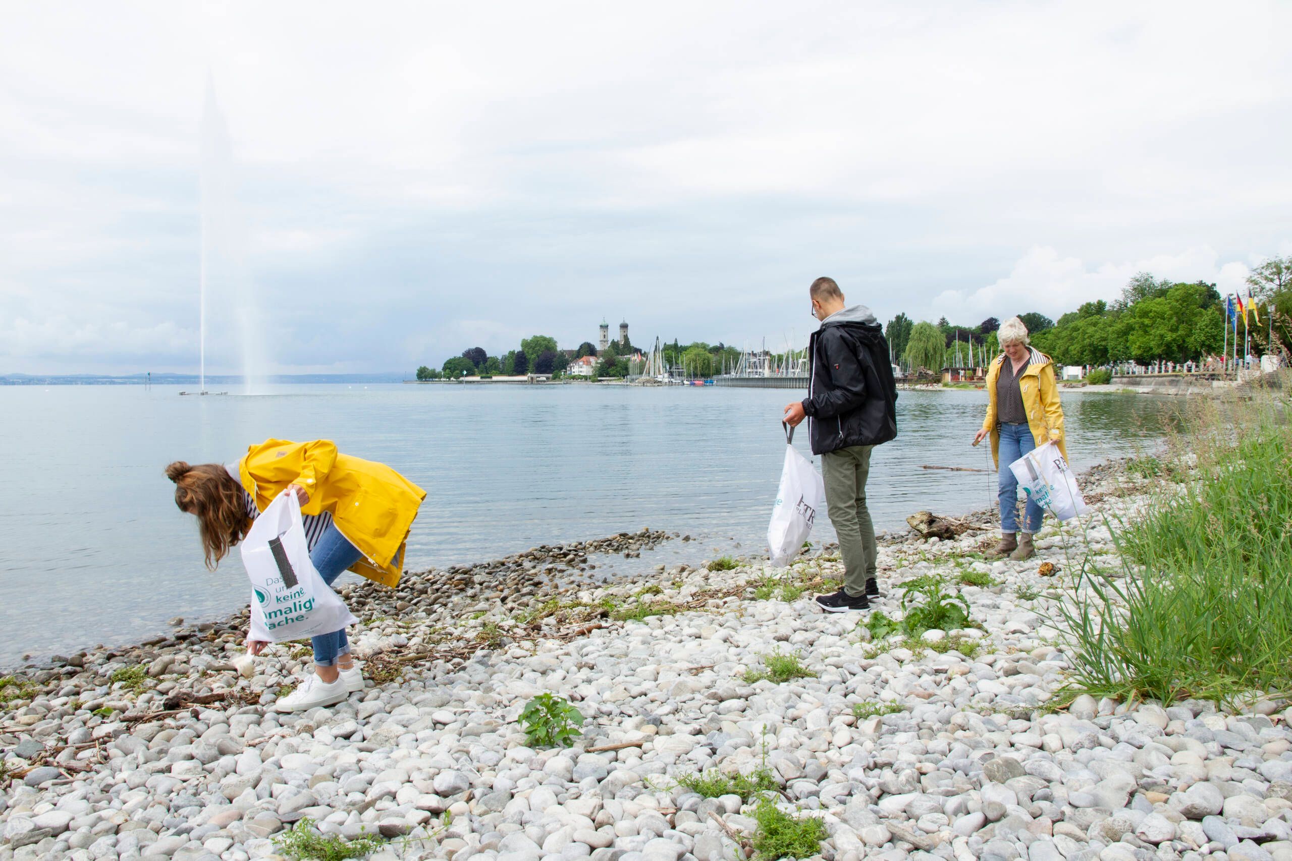 Drei Personen die am Seeufer vom Bodensee Müll einsammeln. Im Hintergrund ist der See zu sehen.