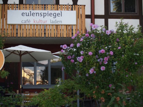 Außenansicht Restaurant und Café Eulenspiegel. Blick auf Terrasse mit blühendem Hibiskus im Vordergrund.