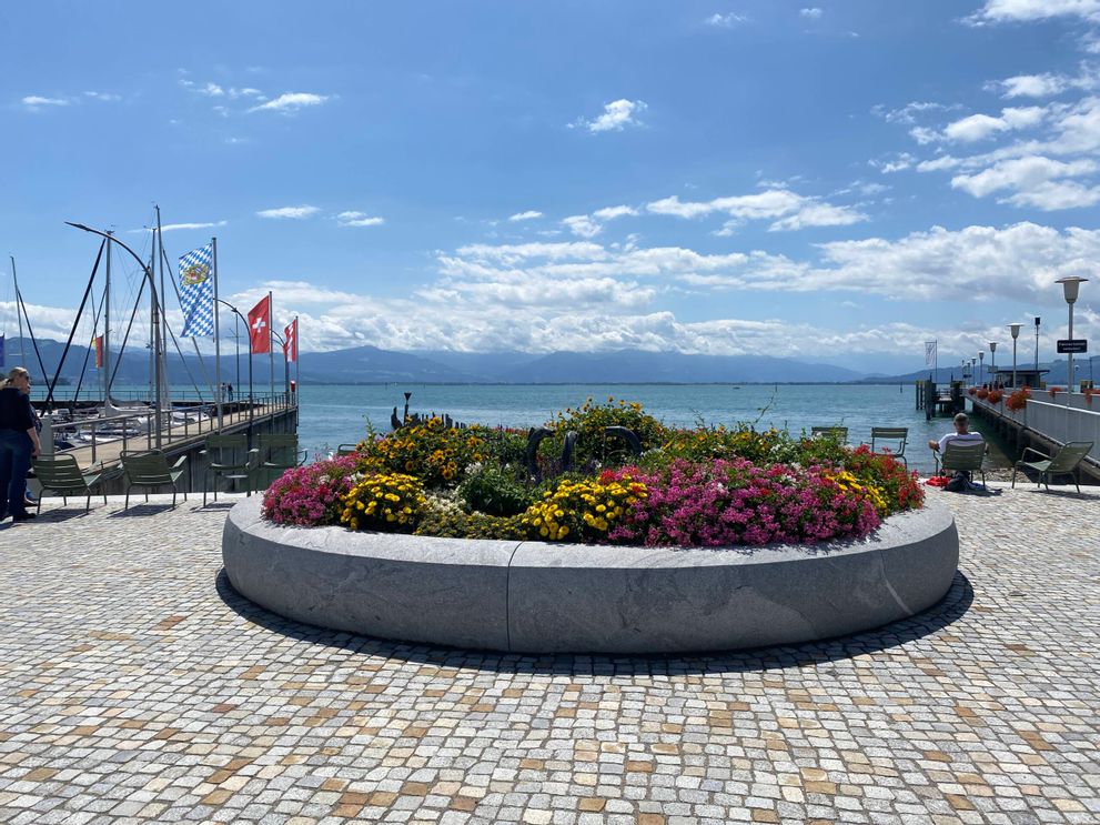 Bepflanzter Brunnen auf der Halbinsel in Wasserburg am Bodensee. Im Hintergrund ist der Bodensee sowie die Bergwelt zu sehen.