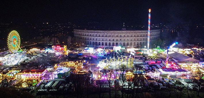 Langzeitbelichtung des Nürnberger Herbstvolksfests bei Nacht mit einem leuchtenden Kettenkarussell und einem Riesenrad, das Lichtstreifen erzeugt, vor der Kulisse der Kongresshalle.