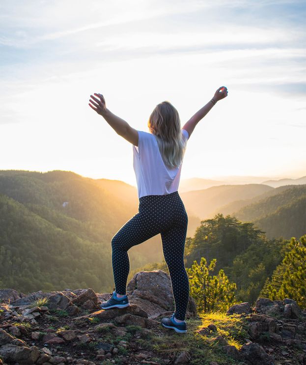 Frau steht auf einem Berg mit ausgebreiteten Armen und blickt über eine Landschaft – Symbol für Gesundheit, Bewegung und Lebensqualität.