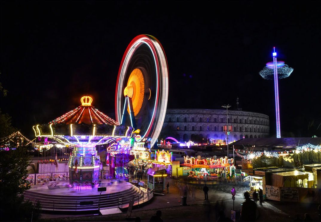 Des Nürnberger Herbstvolksfests bei Nacht mit einem leuchtenden Kettenkarussell und einem Riesenrad, das Lichtstreifen erzeugt, vor der Kulisse der Kongresshalle.