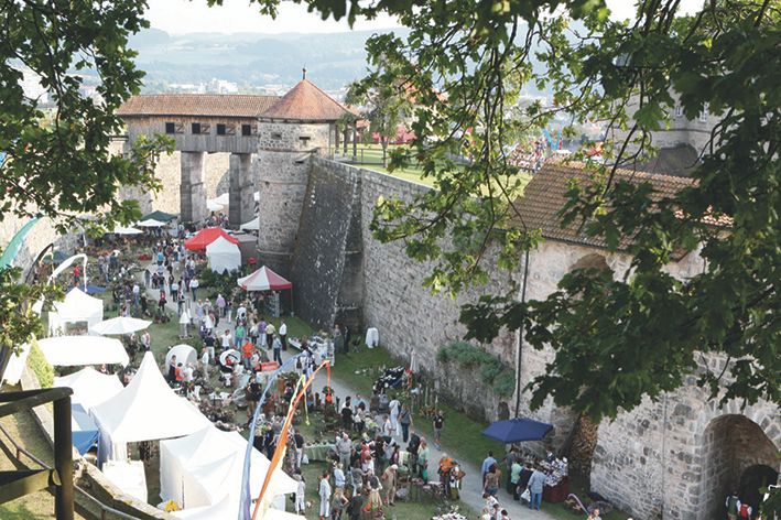Luftaufnahme der Rosenmesse Kronach auf der Festung Rosenberg. Zahlreiche Besucher schlendern zwischen den Marktständen mit weißen und roten Zelten entlang der historischen Festungsmauern unterhalb eines Wehgangs.