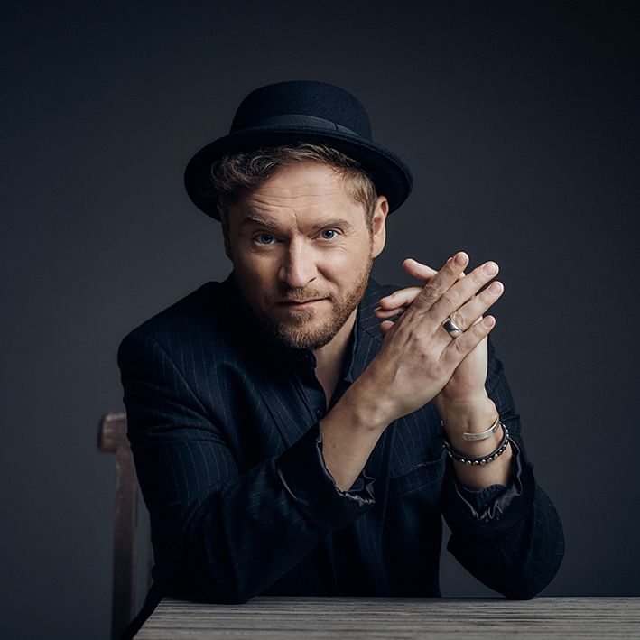 Abschlußkonzert beim Sternenfestival in Burgebrach: Sänger Johannes Oerding in einem Porträtfoto. Er trägt einen schwarzen Hut und ein dunkles Sakko, sitzt an einem Holztisch und blickt mit gefalteten Händen direkt in die Kamera.