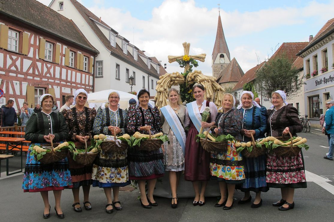 Eine Gruppe von Frauen in fränkischer Tracht und die bayerische Meerrettichkönigin posieren auf dem Baiersdorfer Krenmarkt mit Körben voller Meerrettichwurzeln vor einer großen Skulptur aus Kren.