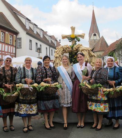 Eine Gruppe von Frauen in fränkischer Tracht und die bayerische Meerrettichkönigin posieren auf dem Baiersdorfer Krenmarkt mit Körben voller Meerrettichwurzeln vor einer großen Skulptur aus Kren.