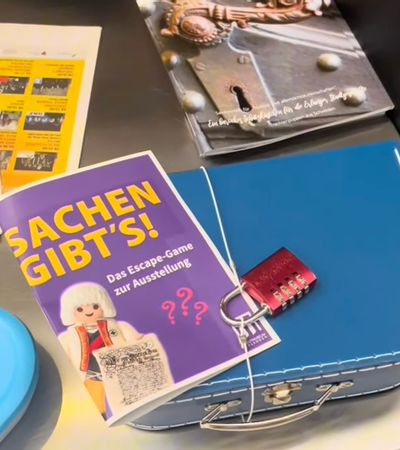 Ein alter, verschlossener Reisekoffer liegt neben einem Rätselheft und einer Lupe, als Szene für das Escape Spiel im Stadtmuseum Erlangen.