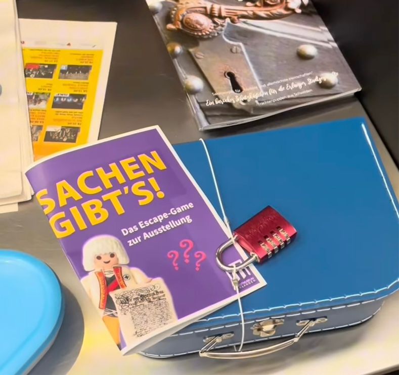 Ein alter, verschlossener Reisekoffer liegt neben einem Rätselheft und einer Lupe, als Szene für das Escape Spiel im Stadtmuseum Erlangen.