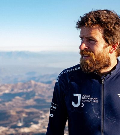 Jonas Deichmann beim Fernweh Festival 2025 in Erlangen. Es zeigt eine Nahaufnahme des Sportlers mit Fahrradhelm und Rucksack vor einer dramatischen Bergkulisse im Abendlicht.