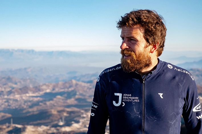 Jonas Deichmann beim Fernweh Festival 2025 in Erlangen. Es zeigt eine Nahaufnahme des Sportlers mit Fahrradhelm und Rucksack vor einer dramatischen Bergkulisse im Abendlicht.